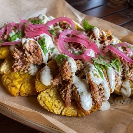 Twice-fried green plantains topped with mojo pork, garlic crema, cilantro and pickled red onion — Loaded Tostones at Two Sisters Kitchen, Flint MI